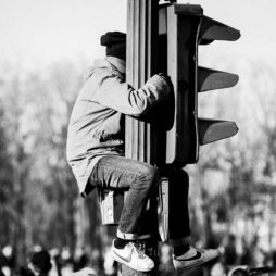 A person wearing a jacket, hat, and sneakers climbs and clings to a traffic light pole, with a blurry crowd and trees visible in the background. The image is black and white.