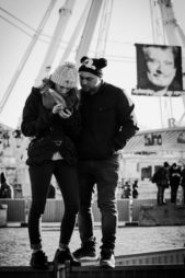 A woman and man, both wearing winter hats, stand close together looking at a phone. Behind them is a large ferris wheel and a banner with a face on it. The photo is in black and white.