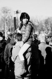A woman wearing a winter coat and fur hat sits on someone's shoulders in a crowd outdoors, looking off to the side. The image is in black and white.