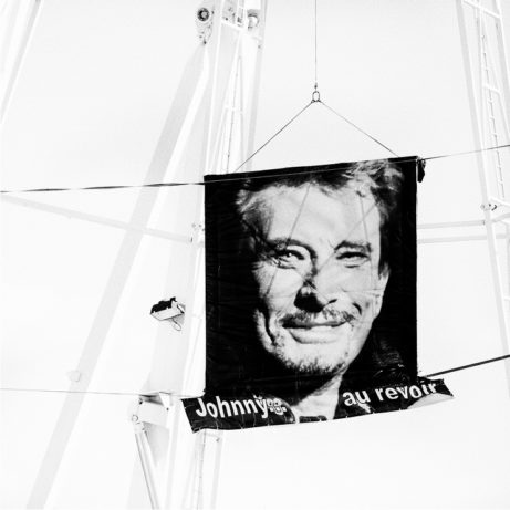 A black and white photo of a large banner showing a man's face, with text at the bottom reading "Johnny au revoir." The banner is suspended outdoors among tall metal structures.