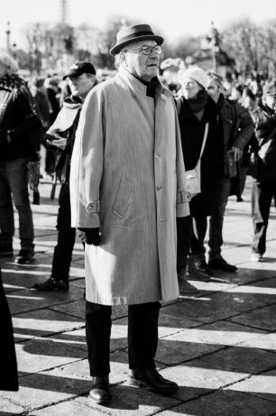 Black and white photo of an older man wearing a light overcoat, hat, and glasses, standing in a crowd outdoors on a sunny day. Other people in coats and hats are visible in the background.