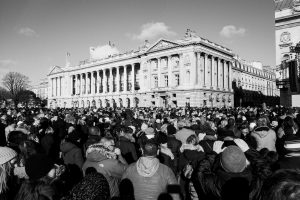 A large crowd gathers in front of a grand, neoclassical building with columns under a sunny sky. People are dressed in winter clothing, and the scene appears lively and busy.