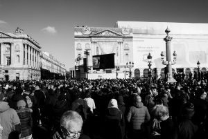 A large crowd gathers in a city square with historic buildings, a fountain, and billboards. People are wearing winter clothing, and a large screen is visible in the background. The image is in black and white.