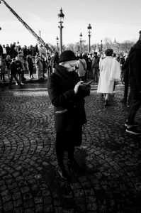 A person in a dark coat and hat stands on a cobblestone street, looking at a phone, while a crowd of people gathers in the background under a clear sky with street lamps visible.