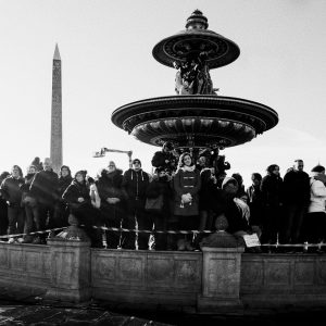A group of people stand around a large ornate fountain in a city square, with an obelisk visible in the background. The people are dressed in winter clothing and the scene is in black and white.