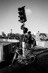 A person with a backpack stands by a traffic light, next to a bicycle locked to the pole. The scene is in black and white, with people and buildings visible in the background under a clear sky.