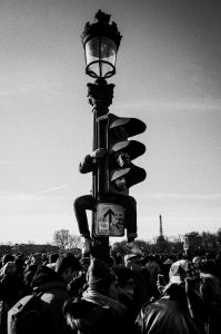 Two people sit on a traffic light pole above a crowded street protest, with many people below. The Eiffel Tower is visible in the background under a clear sky. The image is in black and white.