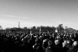 A large crowd gathers outdoors under a clear sky, with the Eiffel Tower in the background and trees without leaves, suggesting a winter day in Paris. The image is in black and white.