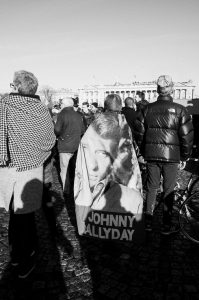 A black-and-white photo of people gathered outdoors; one person has a large cape or flag draped over their shoulders featuring a portrait of Johnny Hallyday and his name printed in bold letters.