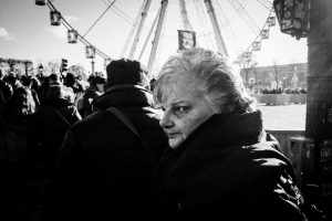 A grayscale photo of a person with short hair and a serious expression looking back at the camera in a crowded outdoor area, with a Ferris wheel visible in the background.