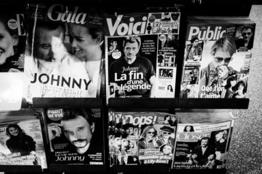 A black-and-white photo of a newsstand displaying various French celebrity magazines, with covers featuring headlines and photos about Johnny Hallyday and other public figures.