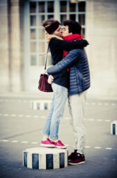 Sandra and Julien share a passionate kiss in an outdoor setting. Sandra, wearing a headband, jacket, and jeans, stands on a striped pedestal, while Julien, sporting a red scarf and jacket, holds her closely. His arm embraces her waist as her legs dangle slightly.