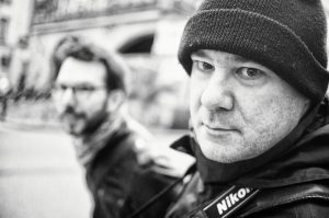 Black-and-white photo of two men; Xavier in the foreground looks directly at the camera, wearing a beanie and carrying a Nikon camera. Fabrice, wearing glasses and a scarf, is out of focus in the background. Urban setting in the background.