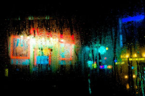 Raindrops on a window blur colorful city lights at night. Neon signs and streetlights create a vibrant mix of reds, blues, and greens against the darkness, casting a dreamy, abstract scene that feels even more surreal when viewed A travers la vitre.