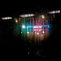 A window covered in water droplets, with blurred neon lights and indistinct shapes visible outside through the wet glass. The neon lights glow in various colors, casting a spirit of mystery and creating a moody atmosphere at night.