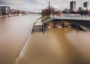 Wide view of the flooded Seine River in Paris. The water has risen over the walkways and sidewalks, submerging steps and extending under bridges while approaching passer traffic near Île aux Cygnes. The Eiffel Tower is visible in the background, and modern buildings line the riverbanks.