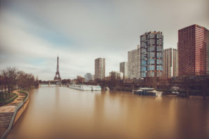 A serene view of the Seine River in Paris, with several modern high-rise buildings lining the Beaugrenelle district on the right bank. The Eiffel Tower is prominently visible in the distance on the left. The overcast sky creates a calm and reflective atmosphere along this stretch of rive gauche.