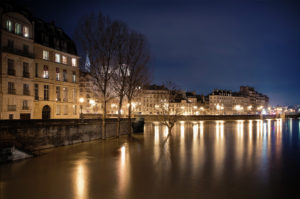 A nighttime view of a cityscape by a river, with buildings illuminated by streetlights reflecting off the water. The sky is dark, and bare trees line the riverbank. The scene exudes a serene and calm atmosphere, as if captured at the bout de la nuit near Deux îles.