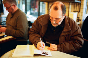 A man with glasses and a mustache, wearing a brown jacket, sits at a table in a café. He is writing in or taking notes from an open book as if preparing for a séance. Another man is seated in the background, also engaged with a book or paper.