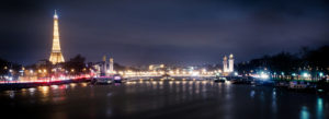 A nighttime panorama of Paris features the illuminated Eiffel Tower on the left and the brille Pont Alexandre-III bridge adorned with lights and golden statues crossing the Seine River, reflecting the city lights. The image captures the serene and enchanting ambiance of the city.