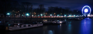 A nighttime riverside scene featuring docked boats in the foreground at Port des Champs-Élysées. In the background, trees line a street illuminated by streetlights. A brightly lit grande roue with a long-exposure effect stands prominently in the distance.