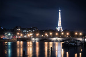 Night view of the Tour Eiffel illuminated, with its reflection shimmering on the Seine River. The Parisian skyline and Pont Alexandre III adorned with streetlights add to the picturesque scene. Boats are docked along Port des Champs-Élysées, contributing to the tranquil ambiance.