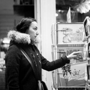 Black and white photo of a woman in a coat with a fur-lined hood, looking at a display with greeting cards. She appears to be browsing or selecting a card in the historic Passage Jouffroy, Paris. The background is a shop or similar indoor setting.