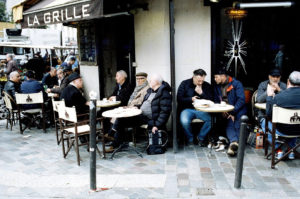 Several men are sitting outside a café named "La Grille" at small round tables. They are engaged in conversation and having drinks. One man is petting a small dog. The café, located near Marché d'Aligre, sits on a cobblestone street with other patrons visible in the background.