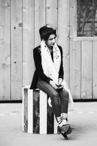 Sandrine Kimperlé sits on a striped cube outdoors, smiling and looking to her left. She is dressed in a blazer, jeans, heeled sandals, and a scarf. A wooden wall is in the background. The image is black and white.