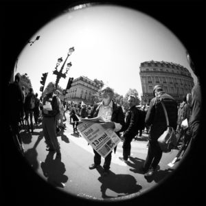 A black-and-white fisheye-lens photograph of a street scene. People are gathered, with a man in the center holding a newspaper that reads "13 mai," possibly referencing "La fête à Macron." Buildings and lamp posts are visible in the background under a clear sky.
