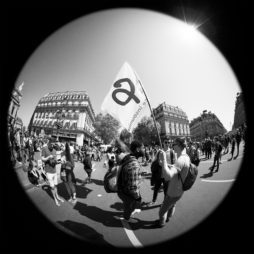 Black and white fisheye photo of a bustling street protest. Two men in the foreground hold a flag with a large "a" symbol, signifying their cause. Surrounding them are other demonstrators from La fête à Macron, some with signs. Buildings and clear sky in the background indicate an urban setting.