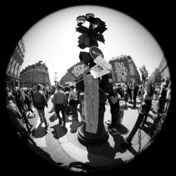 Black and white fisheye lens photo of a crowded street protest. Numerous people gathered around a traffic light, holding signs with messages like "STOP MACRON" and "ARON PRESIDENT." The scene captures the lively energy of "La fête à Macron," illustrating a vibrant urban demonstration.