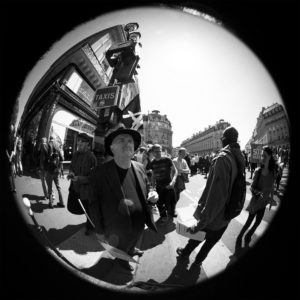 A black and white fisheye photo captures a bustling urban street scene with numerous people walking and standing. One man in the foreground wears a hat and suit. A sign reading "TAXIS" is visible above the crowd, hinting at La fête à Macron, as historic buildings line the street in the background.