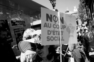 A black-and-white photo showing a woman holding a protest sign that reads, "NON AU COUP D'ÉTAT SOCIAL," during the La fête à Macron. The crowd around her is demonstrating, with other signs and posters visible. The scene appears to be in a city with classical architecture buildings in the background.