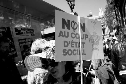 A black-and-white photo showing a woman holding a protest sign that reads, "NON AU COUP D'ÉTAT SOCIAL," during the La fête à Macron. The crowd around her is demonstrating, with other signs and posters visible. The scene appears to be in a city with classical architecture buildings in the background.