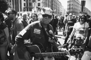 A black and white photo of a street protest. In the foreground, a person is wearing a horned hat that reads "Peuple en Lutte Ensemble," a jacket with various patches including "STOP Macron" and "La fête à Macron," and holding a guitar. Protestors and buildings fill the background.