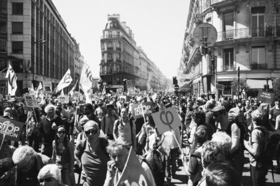 A black and white photo of a large crowd of protestors filling a city street, holding various signs and flags. Tall buildings line both sides of the street, and the atmosphere appears lively and dense. One prominent sign in the center reads "STOP MACRON" amid chants of "La fête à Macron.