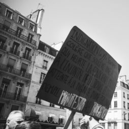 A black and white image of a protest in an urban setting. A prominent cardboard sign held up reads in French: "Indignation (...) Manifestation Indignation." In the background, multi-story buildings with windows and balconies are visible, hinting at the essence of "La fête à Macron.
