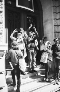 A black-and-white photograph of a group of people standing on the steps of a building. Some are looking up and taking photos with cameras and phones, possibly from a local fête. One person holds a bag and another shields their eyes from the sun with their hand. The building has large doors.