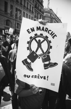 A person at a protest holds a large sign that obscures their face. The sign depicts a gear with a human figure inside and reads "EN MARCHE OU CRÈVE!" in bold text. Other protestors and city buildings can be seen in the background during La fête à Macron. The image is in black and white.