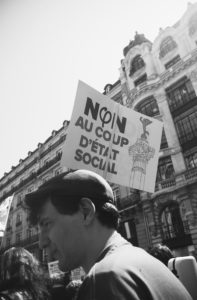 Black and white photo of a street protest. A person in a cap is in the foreground, with a crowd gathered around. A prominent sign reads "Non au coup d'état social" with an illustration of a broken column. In the backdrop are ornate, old buildings, reminiscent of scenes from La fête à Macron protests.