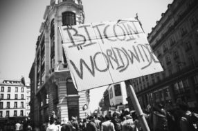 A black and white photo of a city street filled with people. In the foreground, a cardboard sign says "BITCOIN WORLDWIDE" in large, handwritten letters. Tall buildings and a historic structure are visible in the background under a clear sky, reminiscent of fête à Macron gatherings.