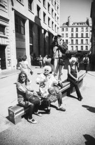 A group of four people, three seated on a low wall and one standing, are in an urban setting. The seated individuals appear to be interacting with each other, perhaps discussing the recent fête à Macron event, while the standing person is taking a photo. The background features tall buildings and other pedestrians.
