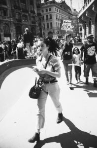 A black-and-white photo captures a city street protest. A woman in the foreground walks purposefully, holding her phone. In the background, children and adults stand and walk; one person holds a sign reading, "STOP MACRON." Historic buildings surround the scene of La Fête à Macron.