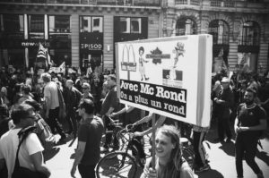 A black and white photo shows a crowd of people marching on a city street. A central figure holds a sign that reads "Avec Mc Rond On a plus un rond" with imagery related to fast food chains. Some marchers are carrying flags and banners proclaiming "La fête à Macron," and bicycles are visible among them.