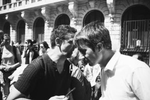 A black-and-white photo shows two men engaged in a close, intense conversation on a crowded street. One man holds a smartphone, perhaps discussing politique, while several other people walk by. The background features a building with arched windows.