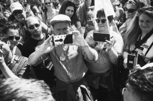 A group of people gathers closely, with many of them holding up smartphones and cameras to take pictures or videos. The crowd appears engaged and focused on something out of view, possibly "La fête à Macron." The image is in black and white, adding a timeless aesthetic.