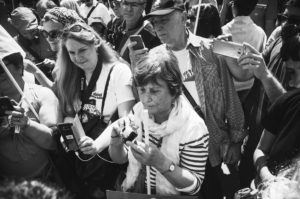 A black and white photograph captures a group of people holding up their smartphones, seemingly taking photos or videos. They appear to be in a busy, outdoor setting, possibly during La fête à Macron, with a mix of emotions visible on their faces.