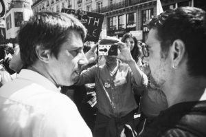 A black-and-white photo of a crowded street protest in France. Two men are in the foreground, facing each other, while a man in the background takes a photo with a camera. People are holding various signs, and buildings are visible in the background.