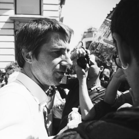 A black and white photo of a man facing to the left, surrounded by people, some holding cameras and taking pictures. The scene appears to be outdoors with buildings in the background. The man is wearing a light-colored shirt and holding a water bottle, reminiscent of La fête à Macron.